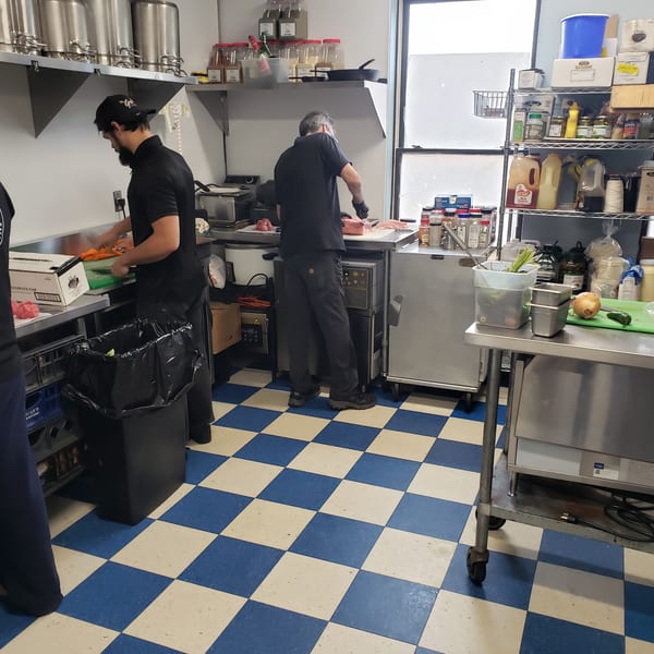 Chefs prepare food in a busy commercial kitchen with blue and white checkered floors.