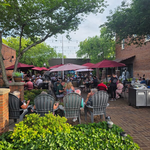 Patrons dine outdoors on a brick patio under red umbrellas and string lights, surrounded by lush greenery.