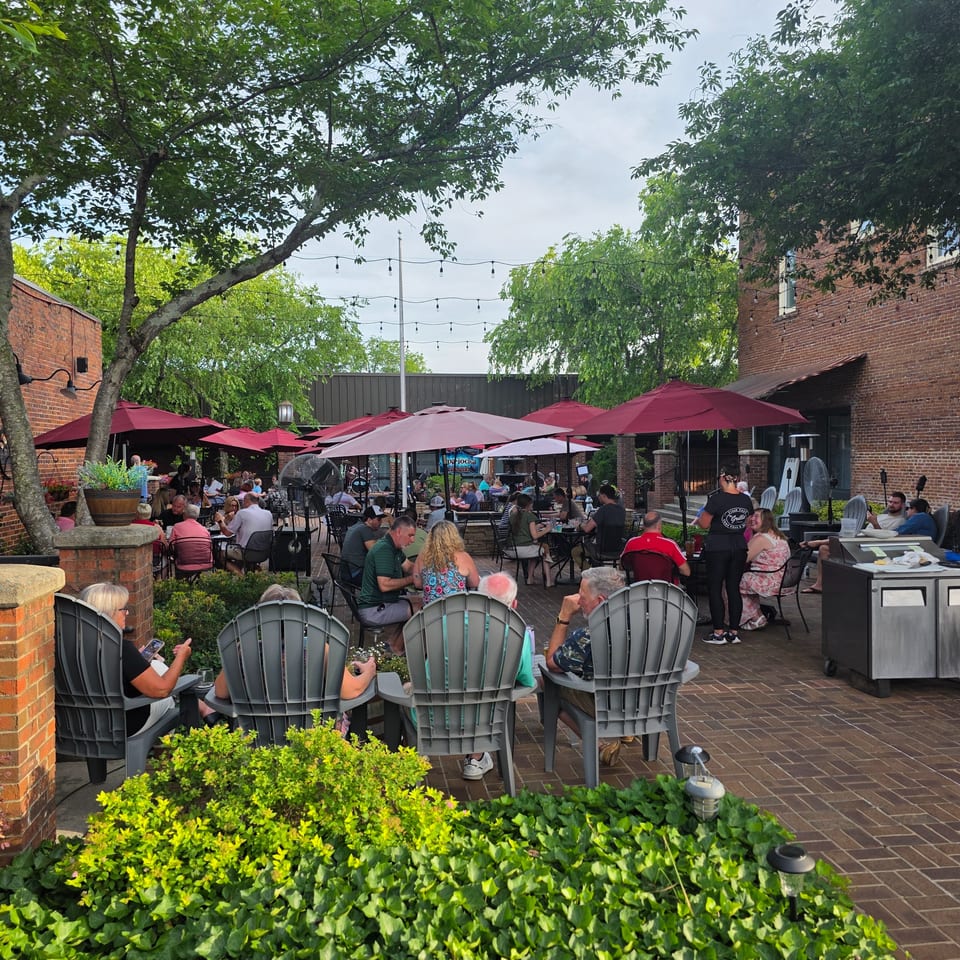 Patrons dine outdoors on a brick patio under red umbrellas and string lights, surrounded by lush greenery.
