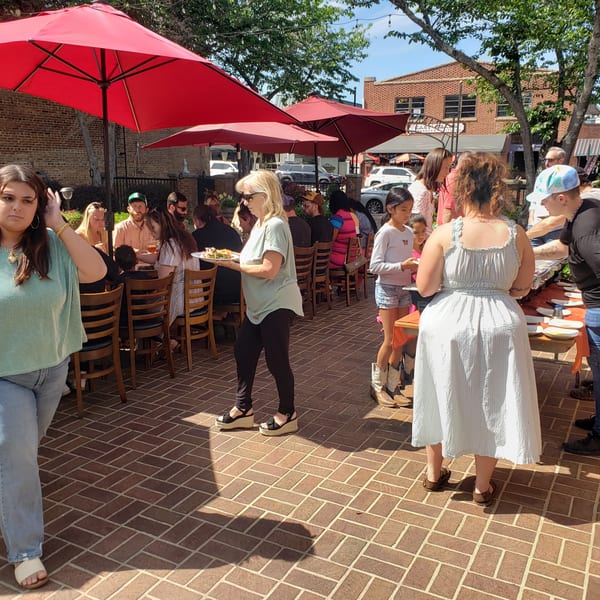 People gather on a sunny outdoor patio with red umbrellas, enjoying food and drinks.