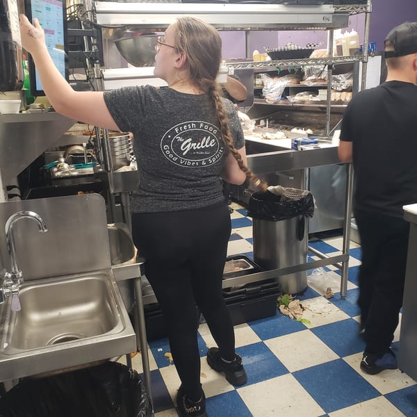 A woman in a "The Grille" shirt uses a touchscreen in a busy commercial kitchen.