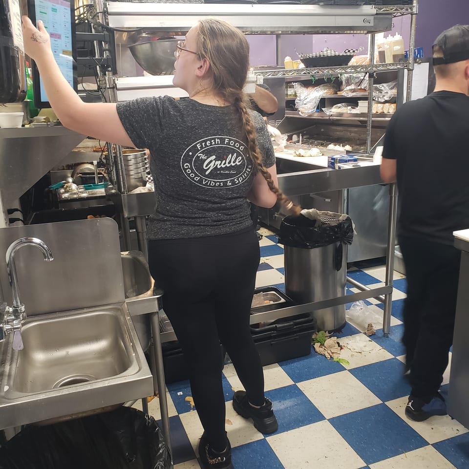 A woman in a "The Grille" shirt uses a touchscreen in a busy commercial kitchen.