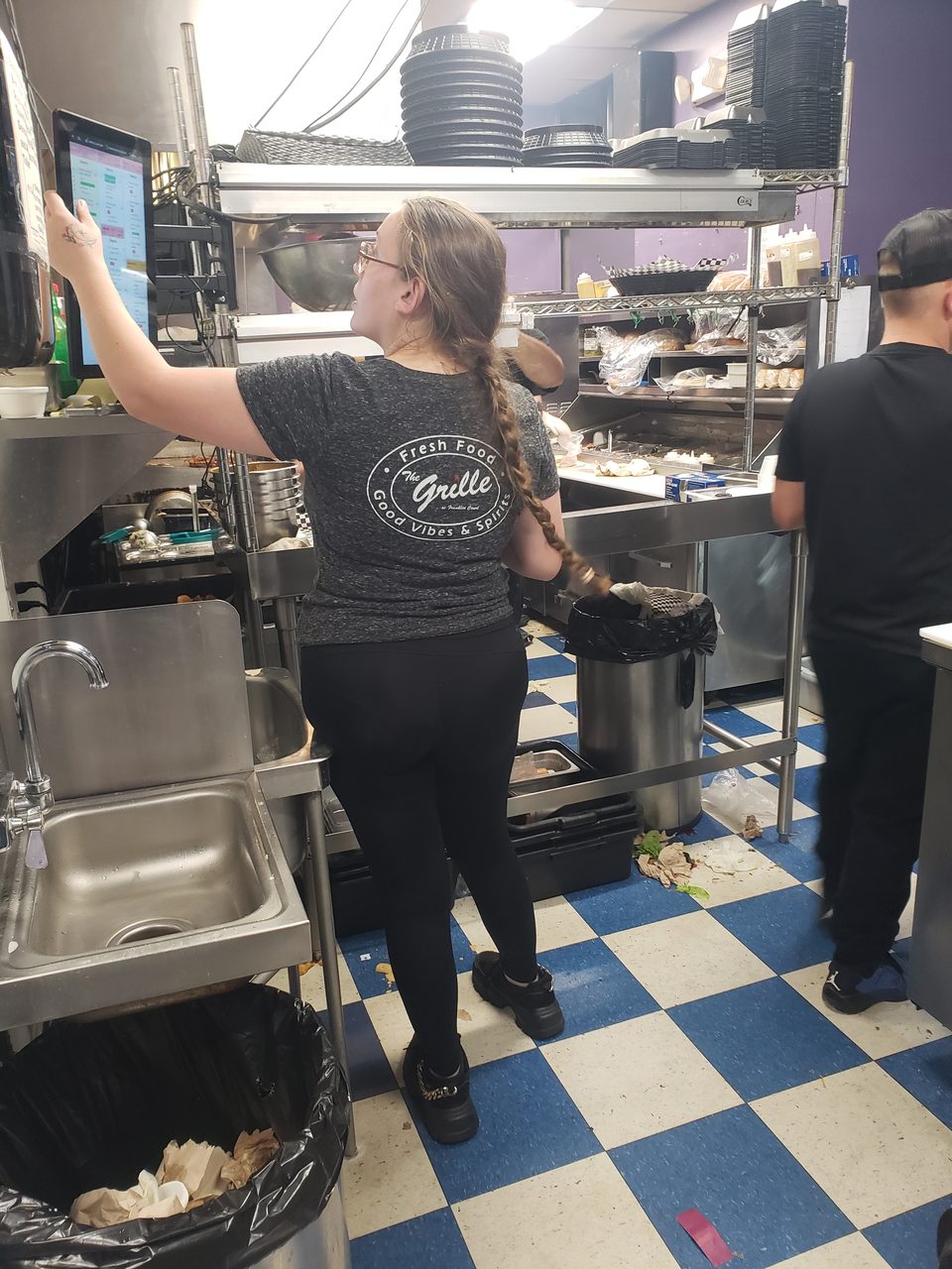 A woman in a "The Grille" shirt uses a touchscreen in a busy commercial kitchen.