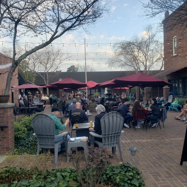 Patrons enjoying an outdoor patio with red umbrellas, string lights, and brick buildings.