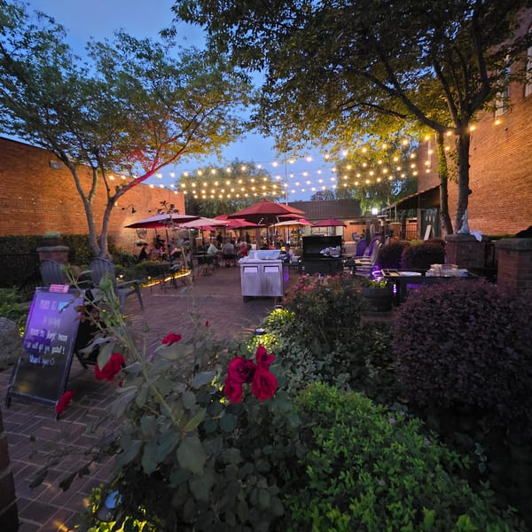 An outdoor restaurant patio at dusk, with string lights, red umbrellas, and lush greenery.