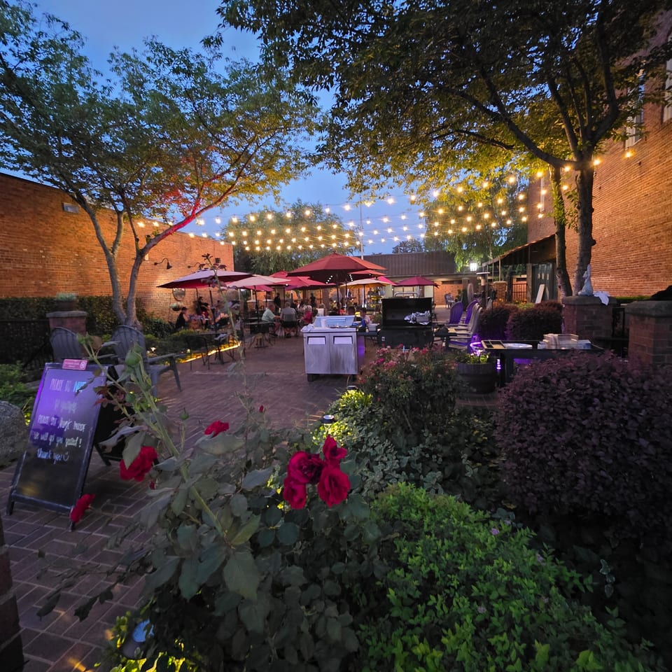 An outdoor restaurant patio at dusk, with string lights, red umbrellas, and lush greenery.