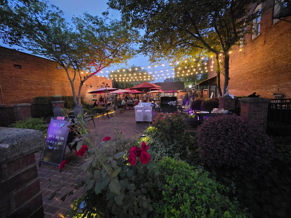 An outdoor restaurant patio at dusk, with string lights, red umbrellas, and lush greenery.
