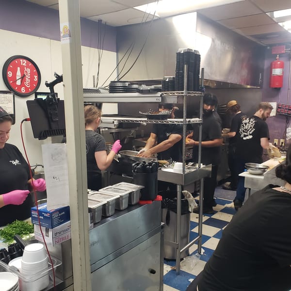 Busy restaurant kitchen with multiple cooks preparing food, stainless steel counters, and a checkered floor.