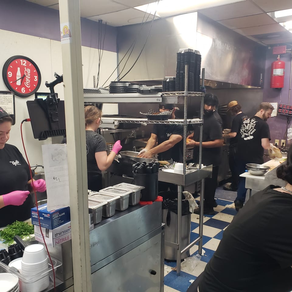 Busy restaurant kitchen with multiple cooks preparing food, stainless steel counters, and a checkered floor.